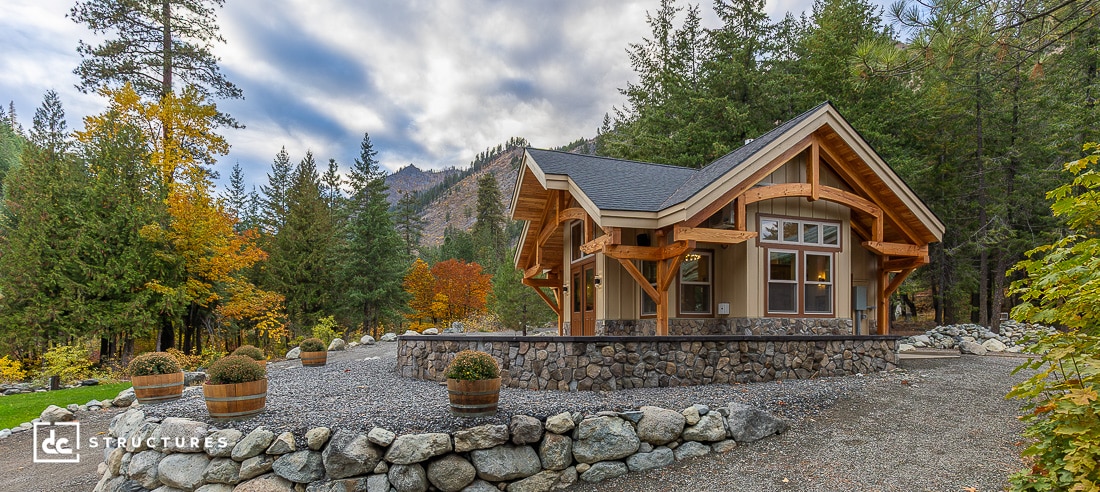 A rustic cabin with large wooden beams and a stone foundation sits among trees and mountains, surrounded by gravel and barrel planters.