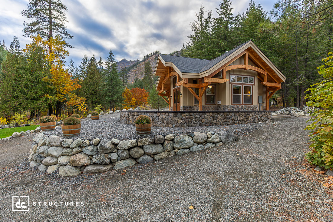 A small modern cabin with timber framing and large windows sits on a gravel lot, surrounded by autumn trees and mountain scenery.