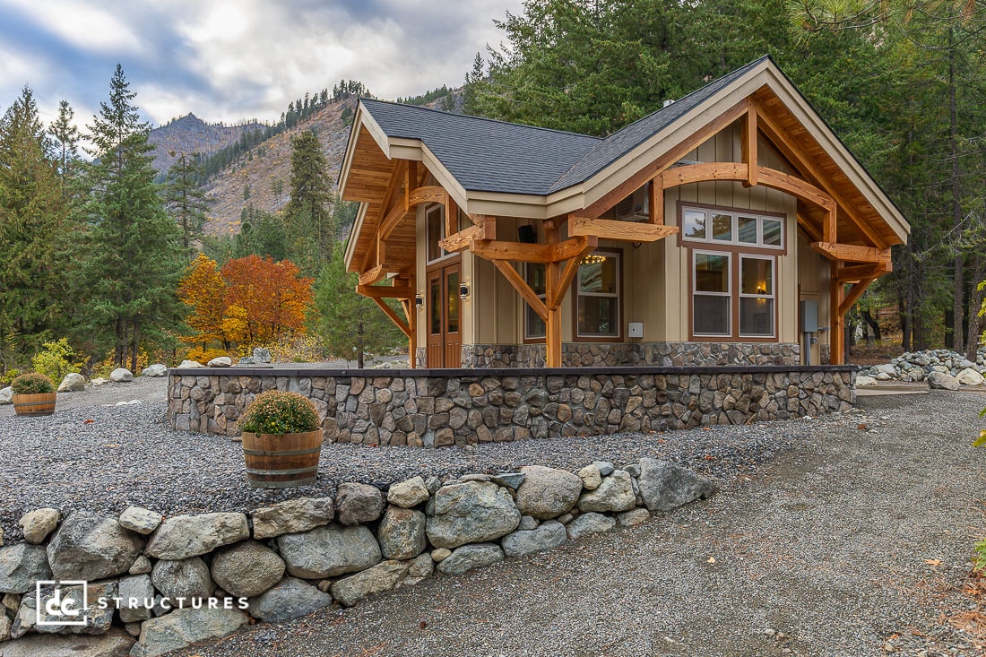 A small modern cabin with stone and wood exterior is nestled among trees and mountains, with gravel, potted plants, and a cloudy sky.