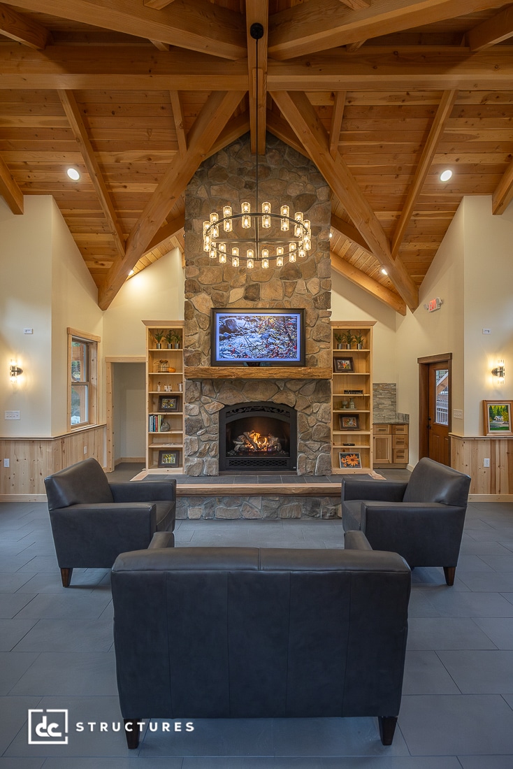 A cozy lounge with dark armchairs surrounds a stone fireplace with a lit fire. Exposed wooden beams and bookshelves add warmth.