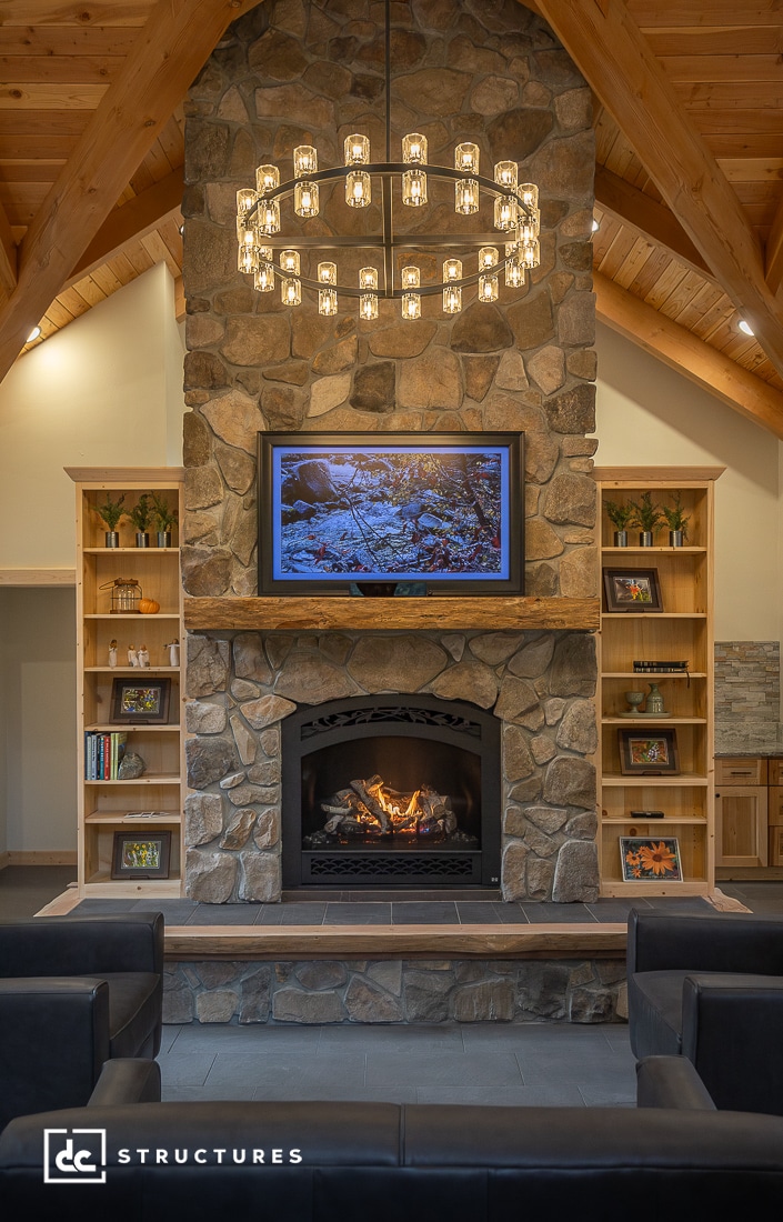 A cozy living room features a stone fireplace with a lit fire, flat-screen TV above, wooden shelves, and a round chandelier.