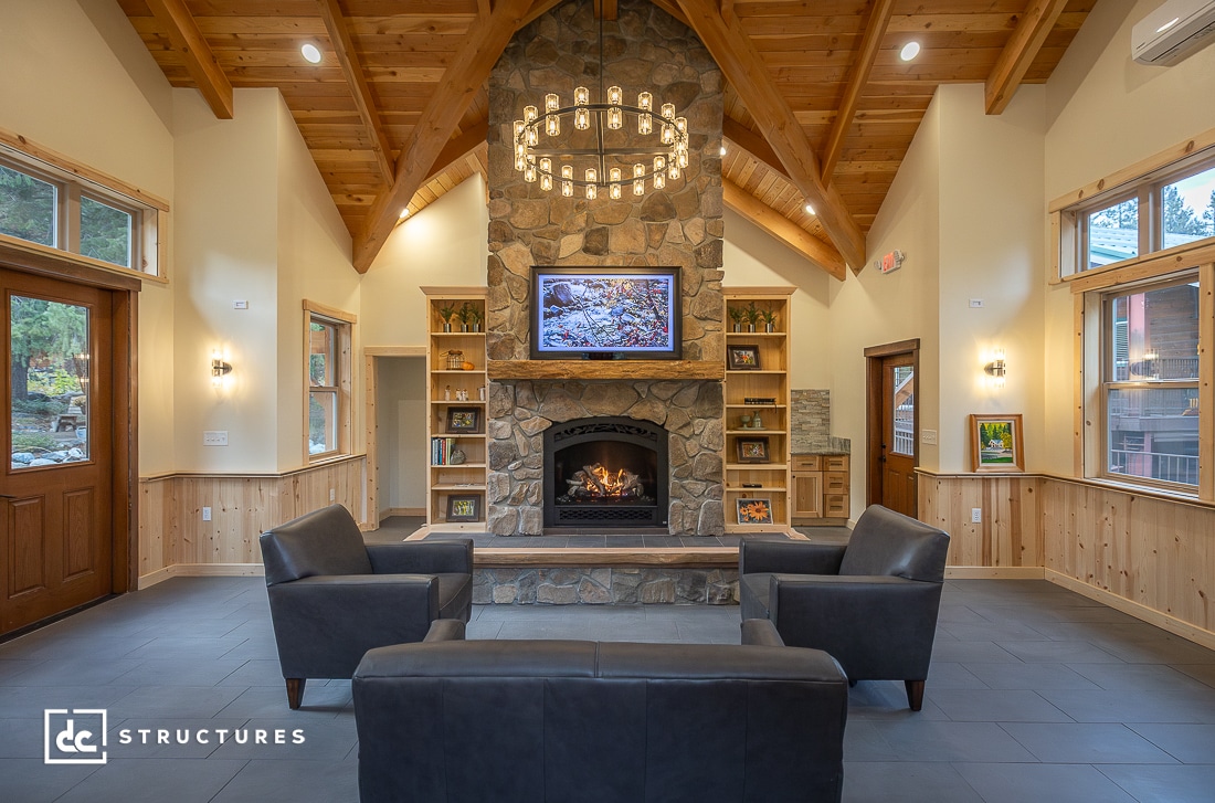 A cozy lodge-style living room with a tall stone fireplace, TV above it, leather armchairs, wood-beamed ceiling, and large windows.