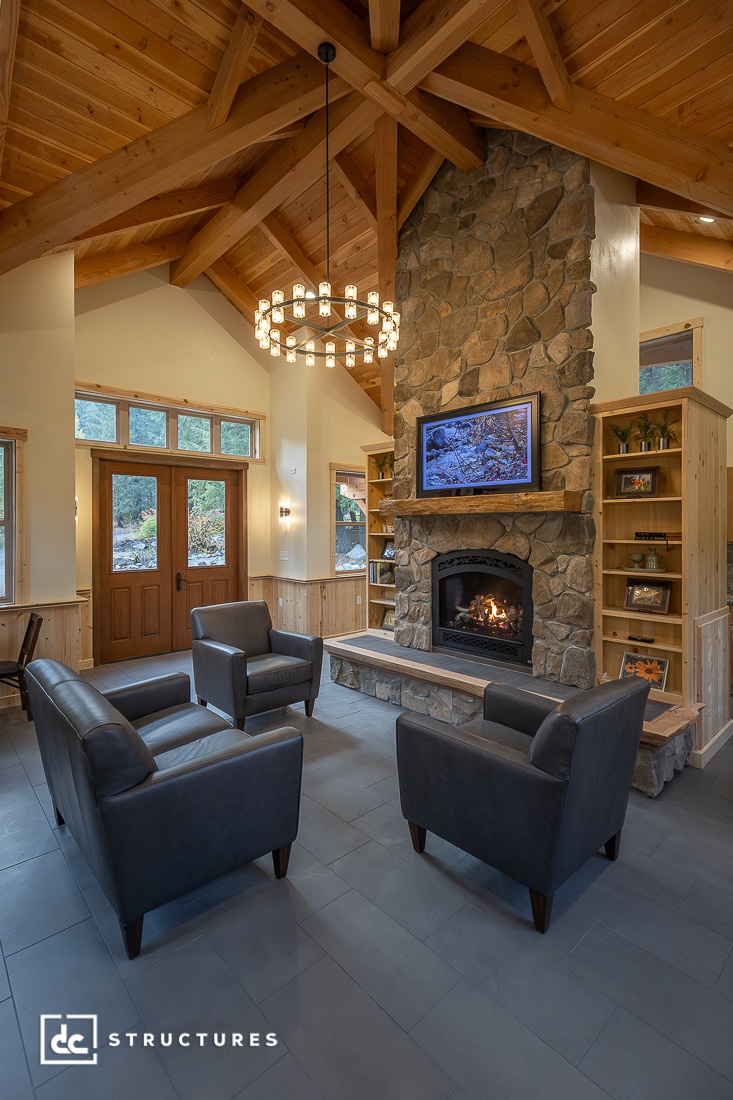 A cozy living room with a tall stone fireplace, a lit fire, two dark armchairs, exposed wooden beams, bookshelves, a chandelier, and large windows with natural light.