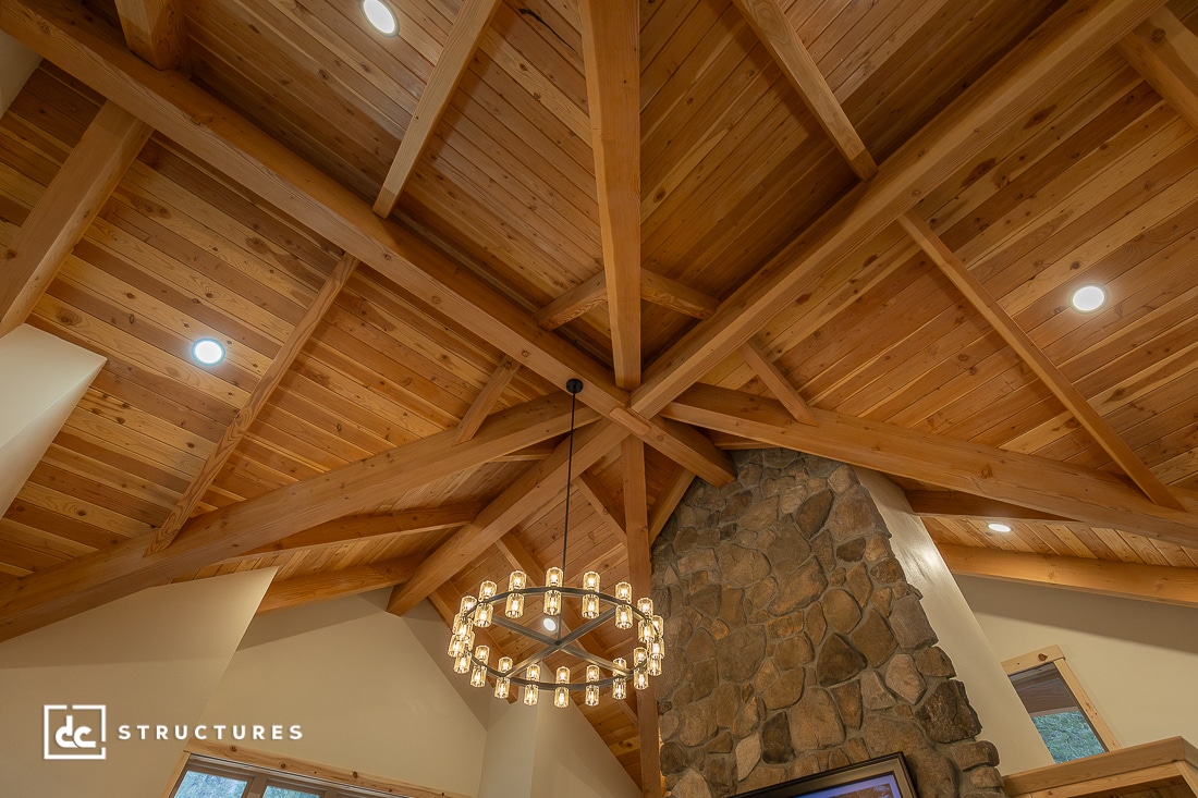 Wooden vaulted ceiling with exposed beams, a circular chandelier, recessed lights, stone fireplace, and partially visible large windows.