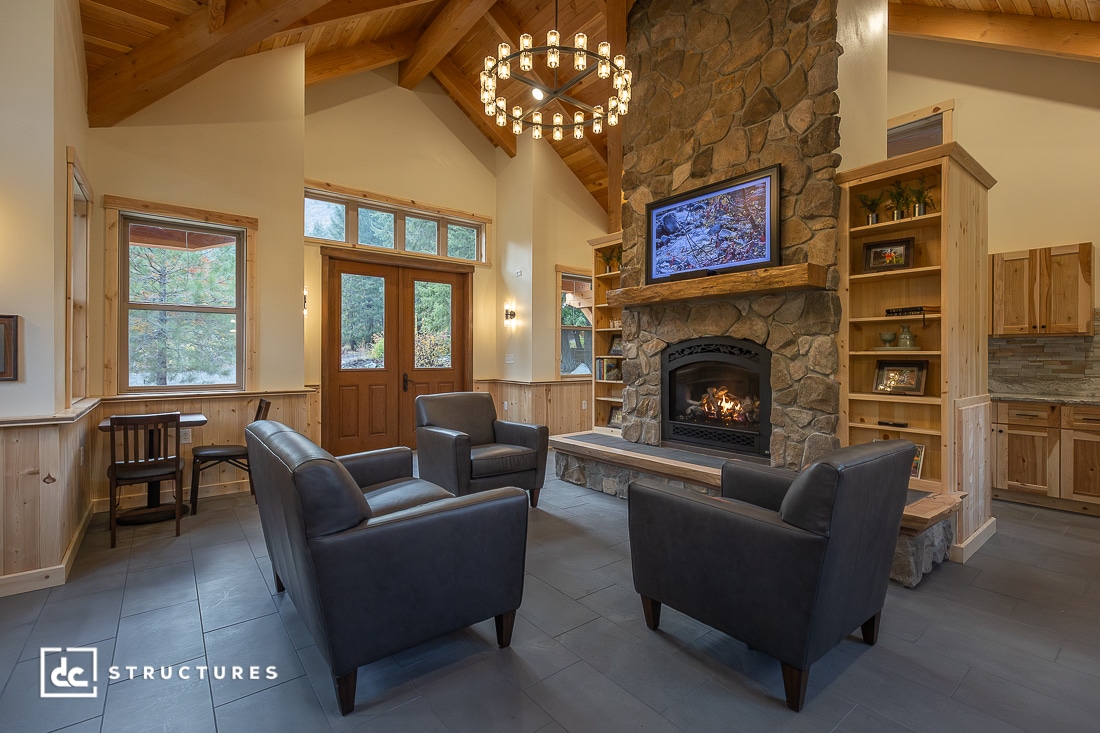A cozy living room with four black armchairs around a stone fireplace, wooden ceiling, shelves, round chandelier, natural light.