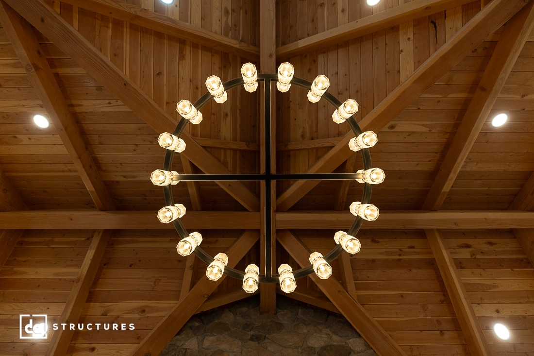 A circular chandelier with exposed bulbs hangs from a high, wooden, vaulted ceiling with visible beams. A stone wall is at the bottom.