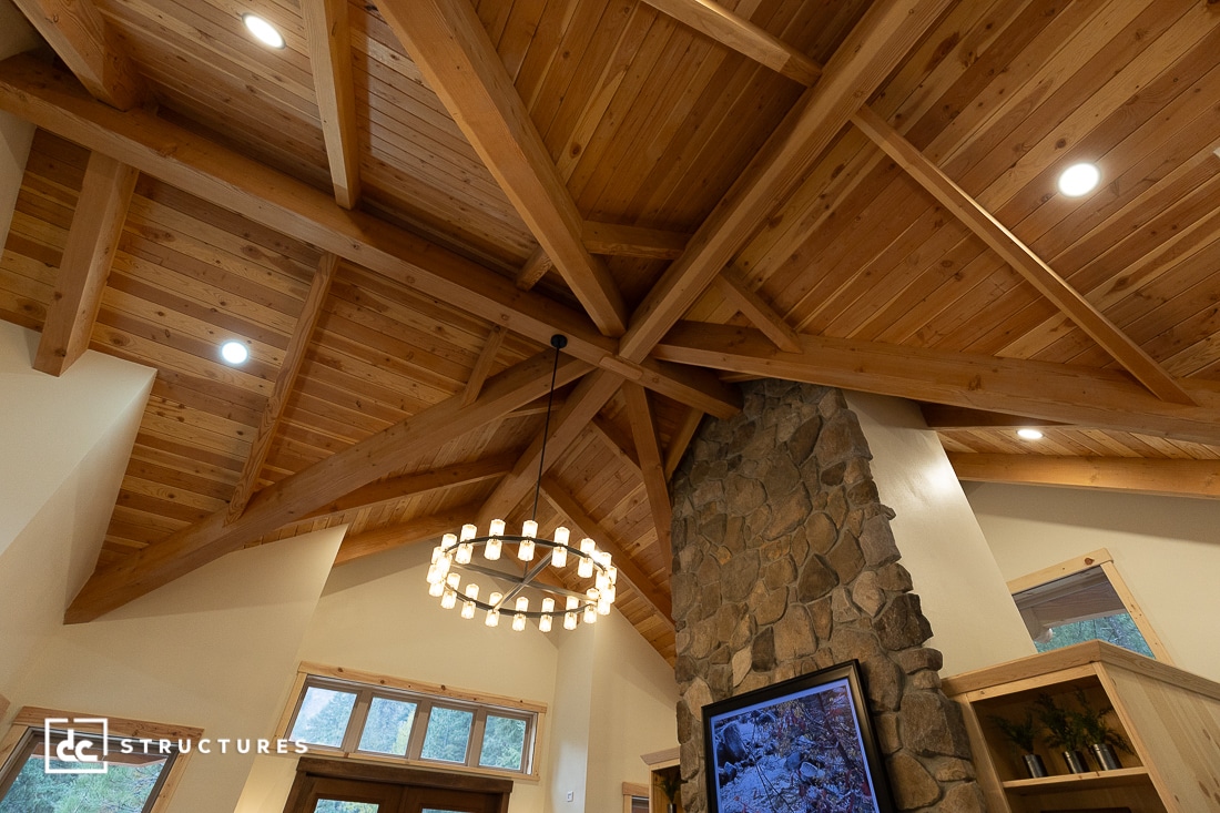 A vaulted wooden ceiling with exposed beams and a modern chandelier hangs over a stone fireplace. Recessed lights and tall windows brighten the rustic, cozy interior.