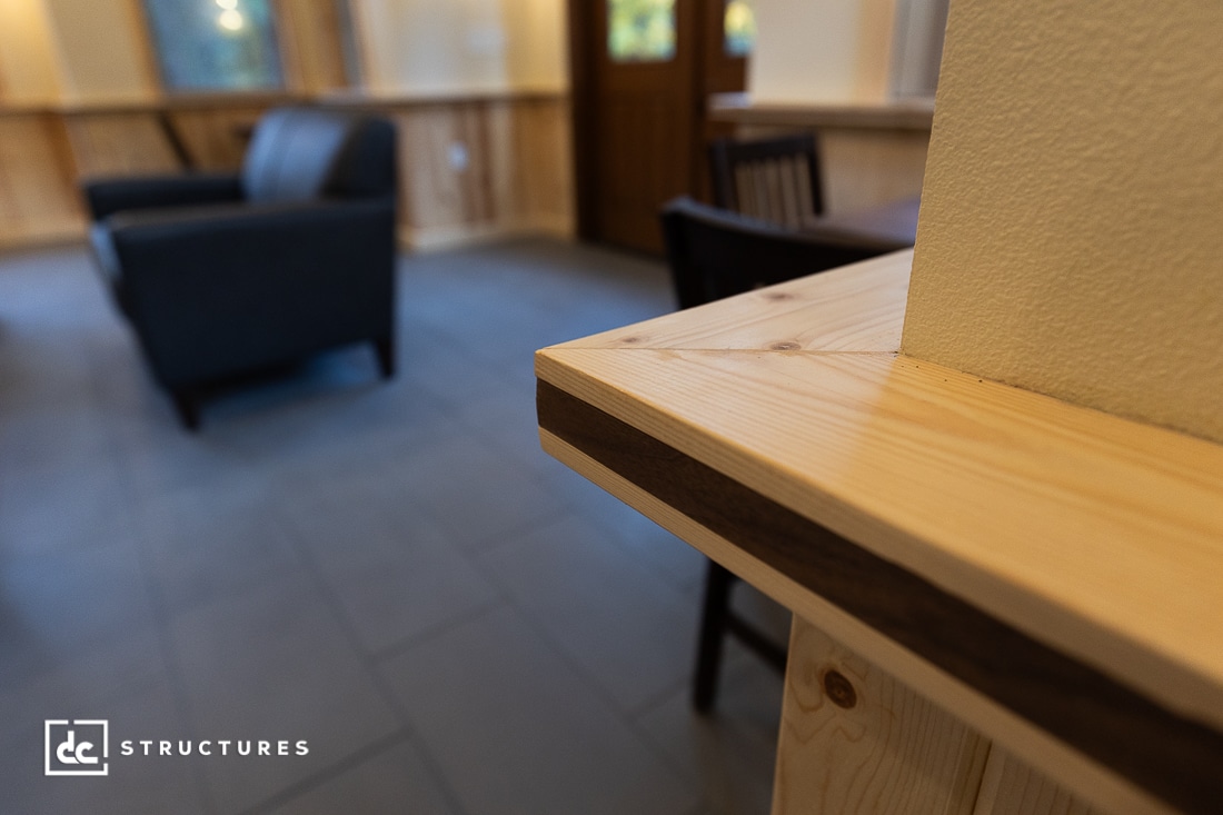 Close-up of a wooden counter with visible grain, set in a room with tile flooring, a sofa, chairs, and wooden wall paneling.