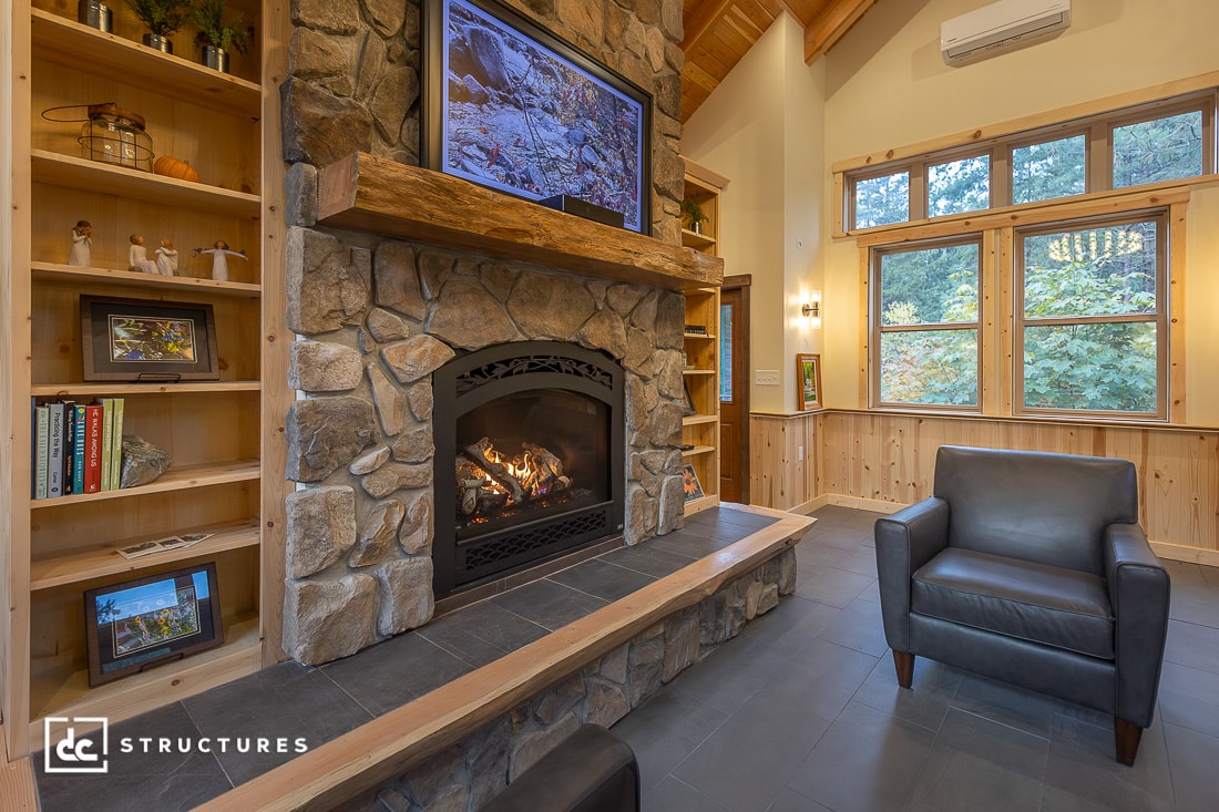 A cozy living room with a stone fireplace, fire burning, TV above, shelves of books and decor, dark armchair, and large windows.