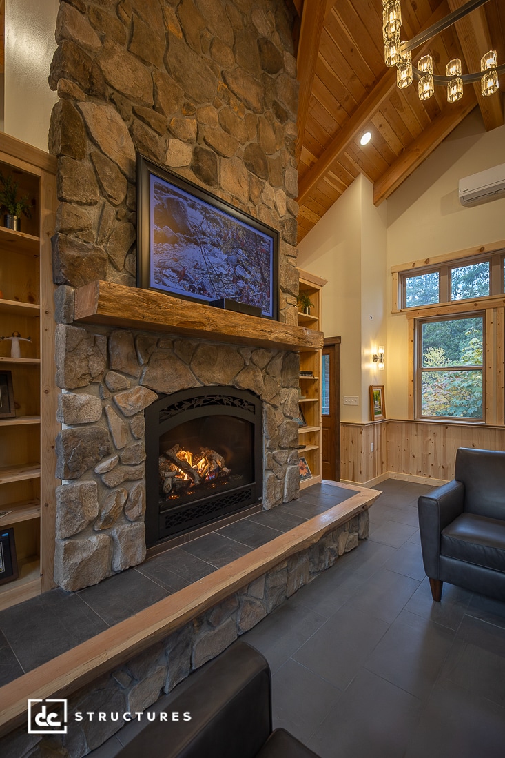 A cozy living room with a large stone fireplace, wooden mantel, wood ceilings, bookshelves, big windows, and black armchairs.