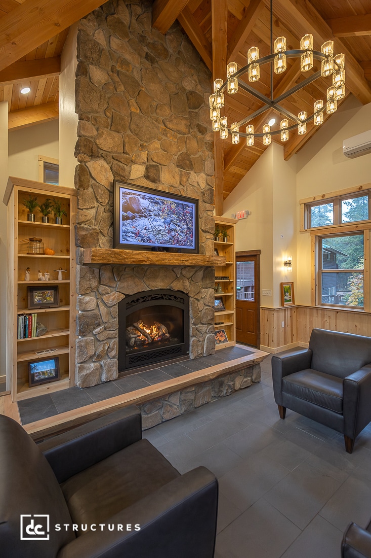 A cozy living room with a tall stone fireplace, wooden mantel, lit fire, dark armchairs, gray floor, shelves, and warm lighting.