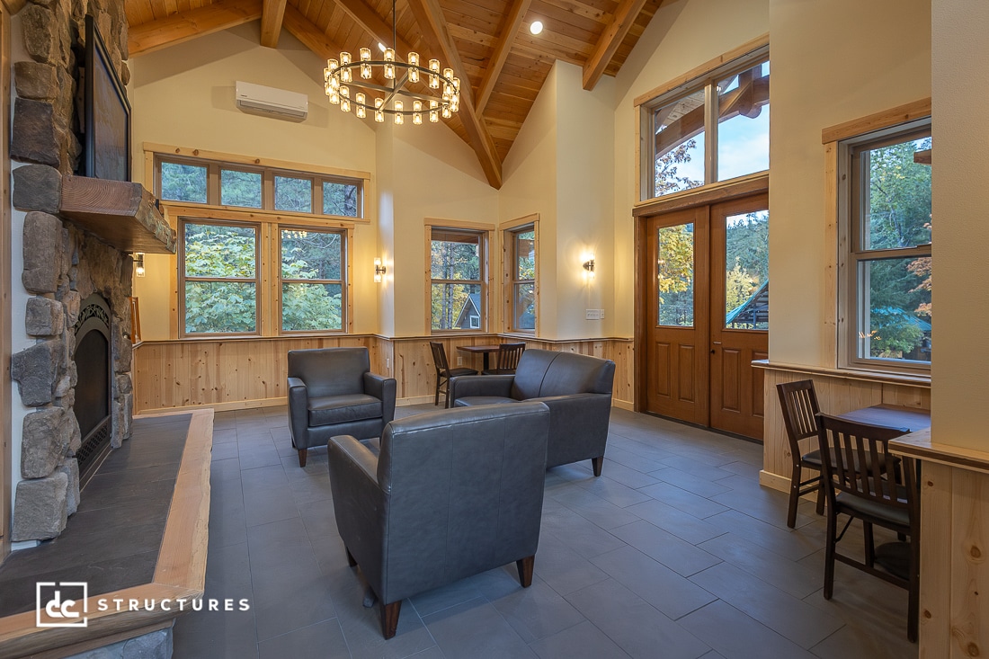 A cozy room with high wood ceilings, large windows, a stone fireplace, leather chairs, small tables, a chandelier, and wood trim. Sunlight streams in through the windows and glass doors, highlighting the warm interior.