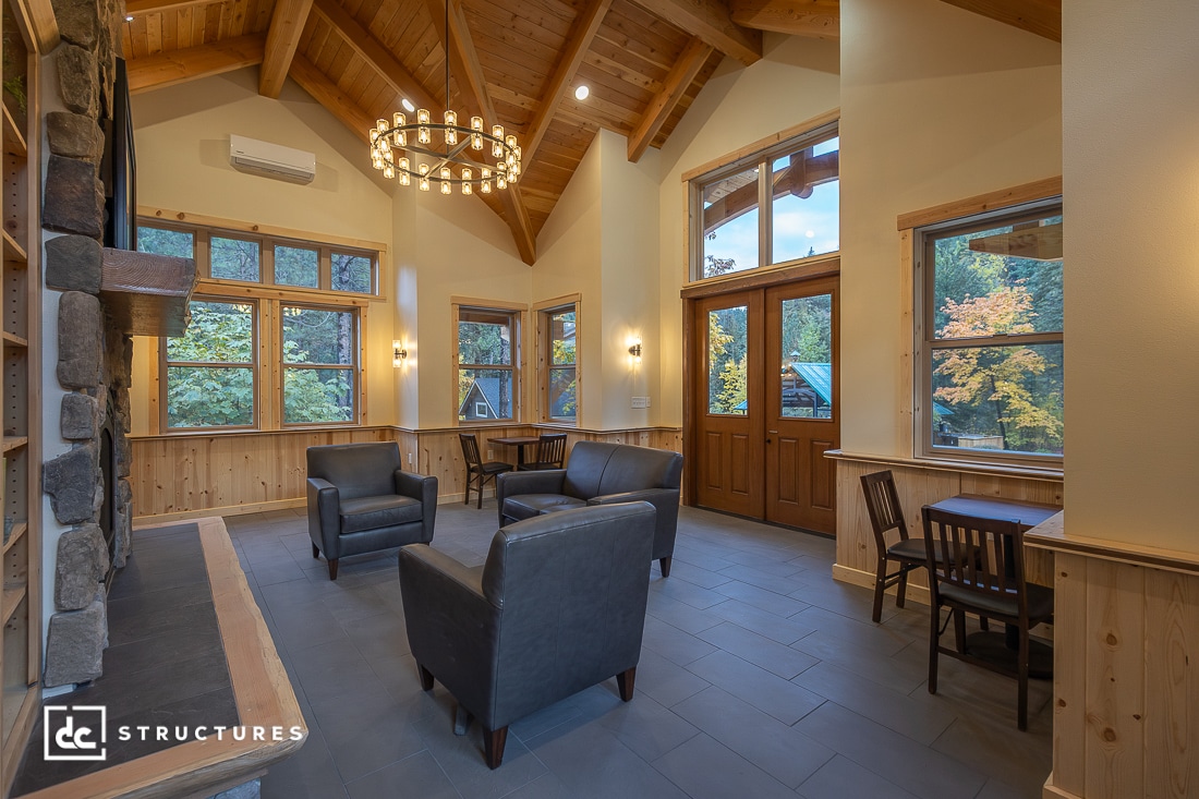 A cozy, high-ceilinged room with large windows, wood accents, and a stone fireplace. Four black armchairs surround a table; trees outside.