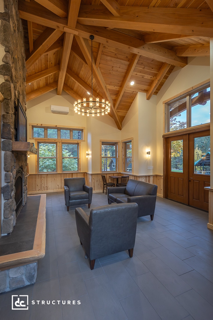A cozy room with a high wood-beam ceiling, stone fireplace, large windows, wooden double door, armchairs, and round chandelier.