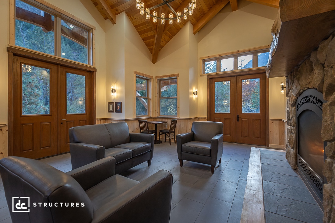 A cozy lodge-style living room with leather chairs, stone fireplace, wooden beams, large windows, and double doors showing forest views.
