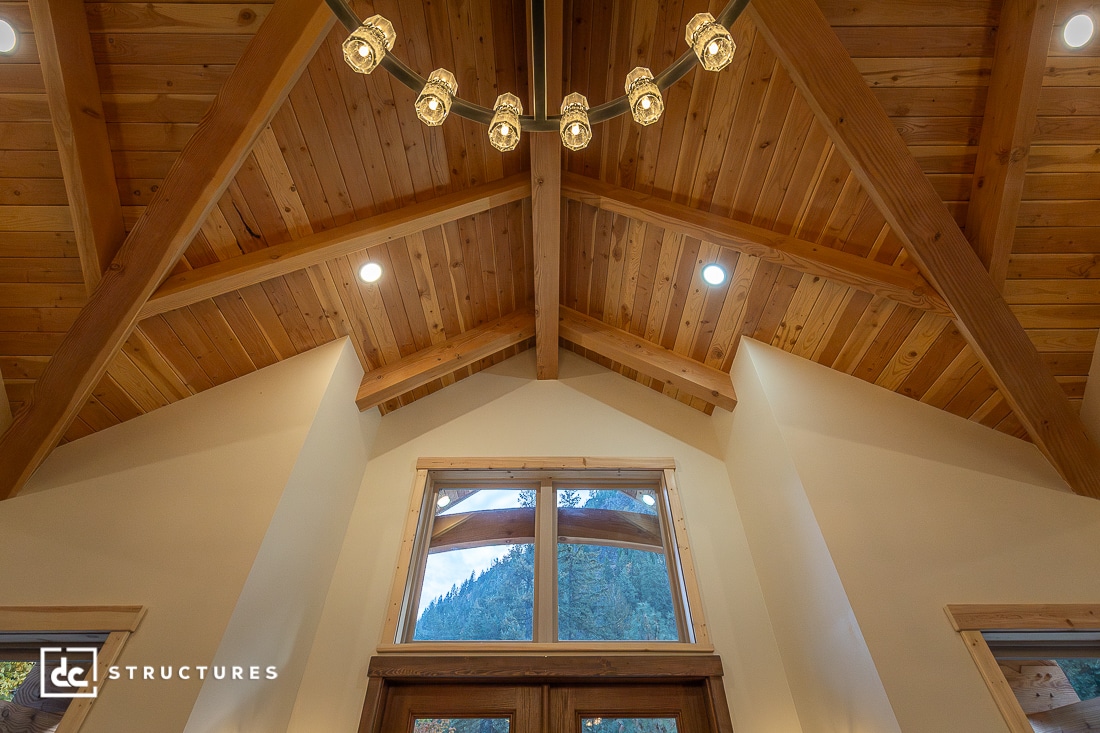 Wood-beamed vaulted ceiling with a modern chandelier, recessed lights, and a large window framing mountains and trees.
