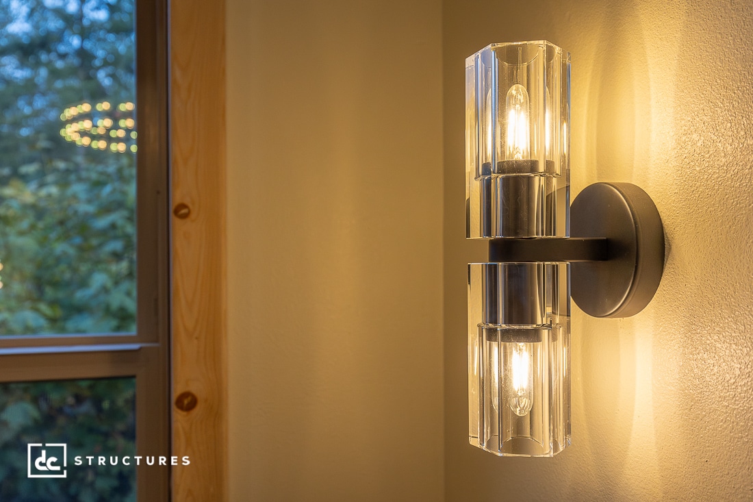 A modern wall sconce with two exposed bulbs and clear glass shades is lit, mounted on a beige wall next to a window with greenery.