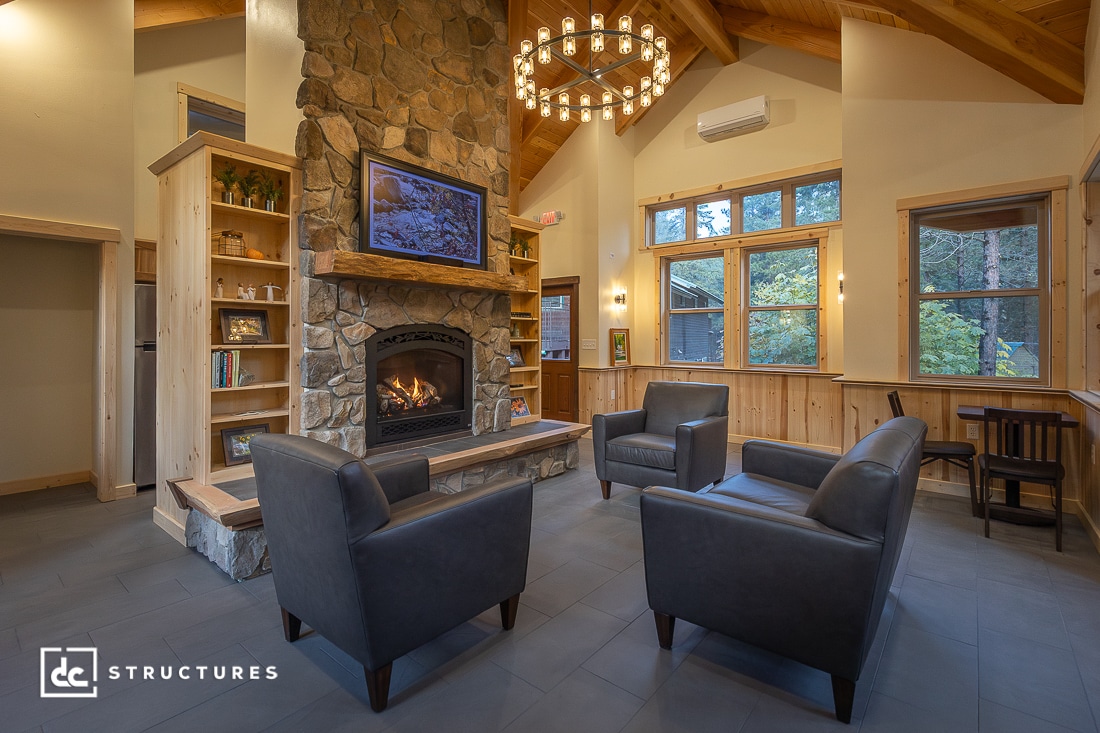 A cozy living room with a stone fireplace, mounted TV, three gray armchairs, wood paneling, large windows, and a chandelier.