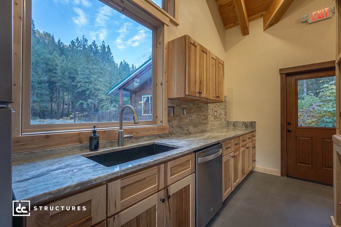 A modern kitchen with wooden cabinets, stone countertops, a stainless steel dishwasher, large window, and rustic natural lighting.