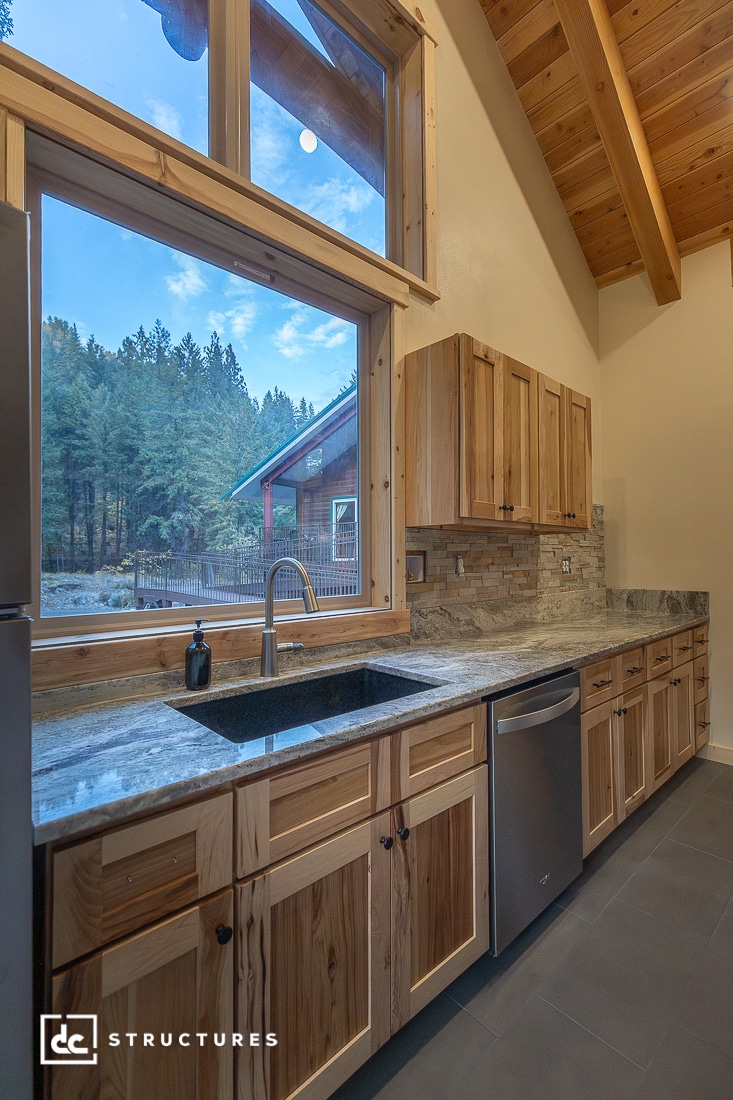 A modern kitchen with natural wood cabinets, granite countertops, stainless steel appliances, and large windows showing a forest view. The ceiling is vaulted with exposed wood beams.