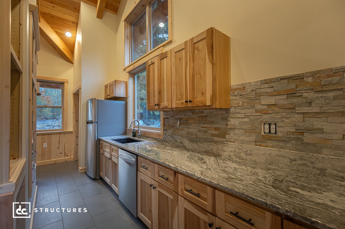 Modern kitchen with natural wood cabinets, granite countertops, stainless appliances, stone backsplash, large windows, high wood-beamed ceilings, and gray tile flooring.
