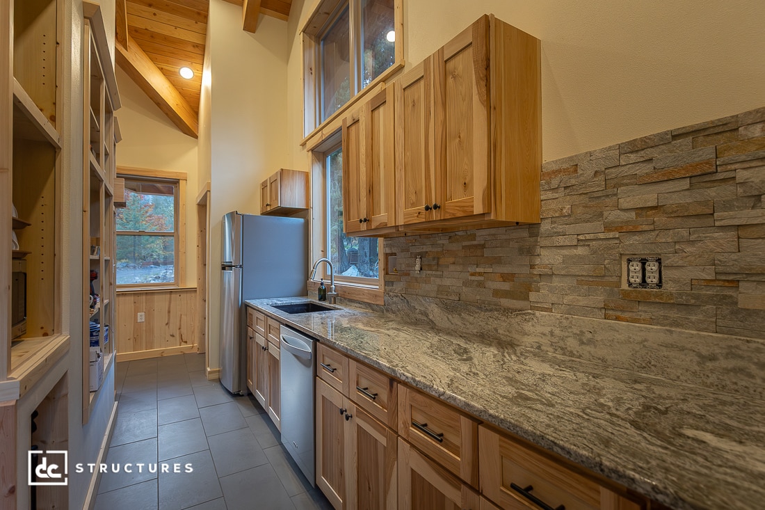 Narrow rustic kitchen with wooden cabinets, gray stone backsplash, marble countertop, stainless appliances, tall windows, and gray tile floor. Warm lighting highlights natural wood features.