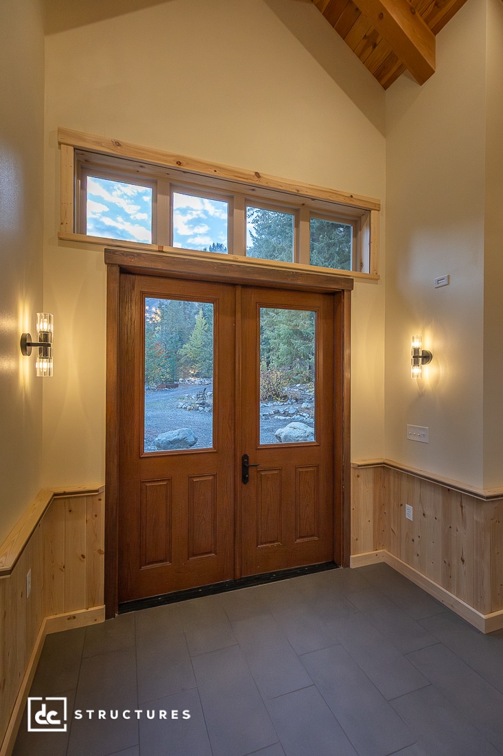 A modern entryway with double wooden doors, glass panels, transom windows, wall sconces, wood trim, and a view of trees outside.