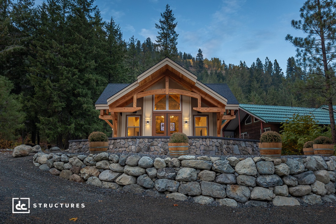 A cozy wooden cabin with large windows and a stone foundation sits surrounded by trees. Four planter barrels line the entry path. Forested hills are visible in the background.