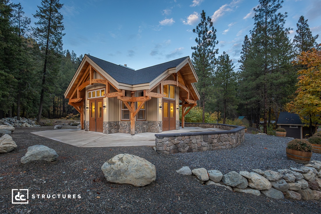 A rustic, wood and stone cabin with large windows is surrounded by trees and rocks, featuring a curved stone patio wall and a forested backdrop under a blue sky.