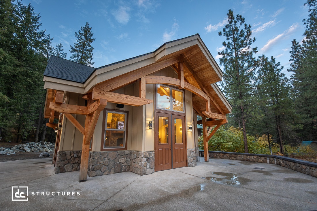 A modern cabin with stone and wood exterior, large wooden beams, and tall windows stands among trees at dusk under a cloudy sky.