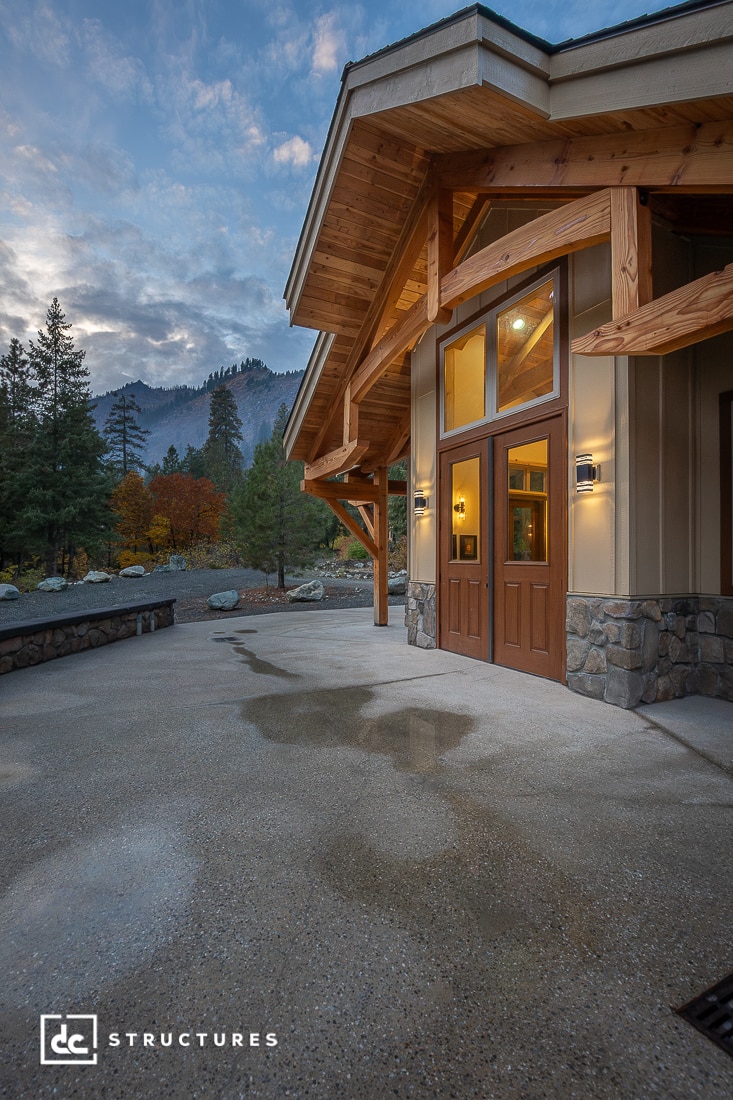 Modern timber-framed building with stone and wood accents, large windows, and covered entryway. Surrounded by trees and mountains at dusk.
