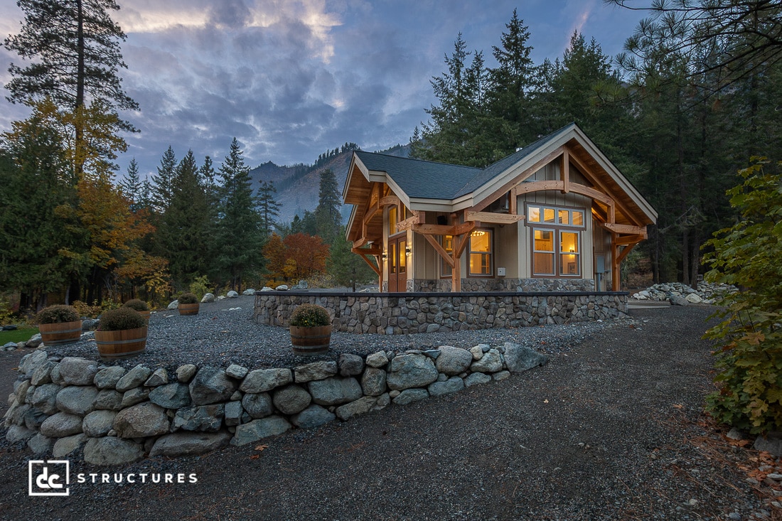 A cozy timber-framed house with large windows on a stone foundation, surrounded by trees and mountains at dusk. Warm interior lighting contrasts with the cool, cloudy evening sky.