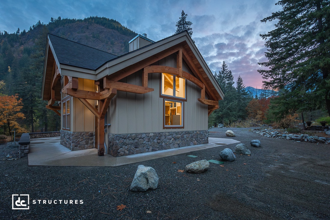 A modern cabin with stone and wood exterior sits in a forested mountain area at dusk, warm light glowing from its windows.