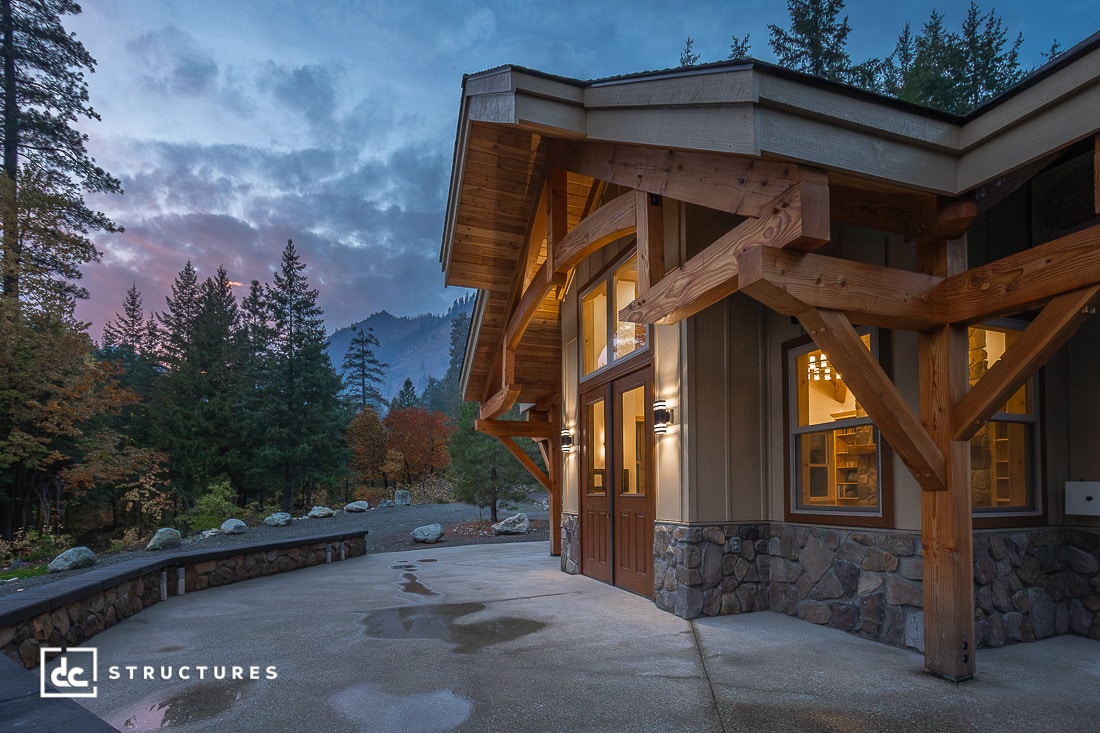 A modern wooden house with large windows and stone accents is surrounded by trees and mountains at sunset. Warm lights glow inside.