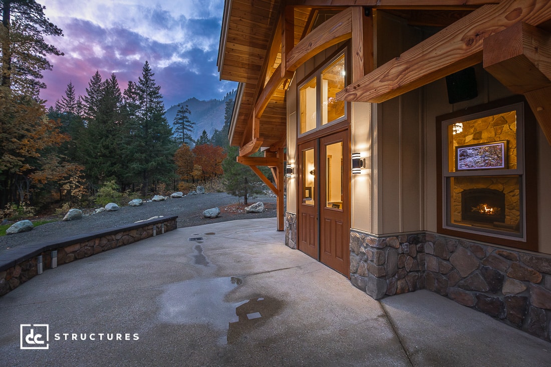 A rustic house with stone and wood exterior by a concrete driveway, surrounded by trees and mountains at sunset. Warm light glows inside.