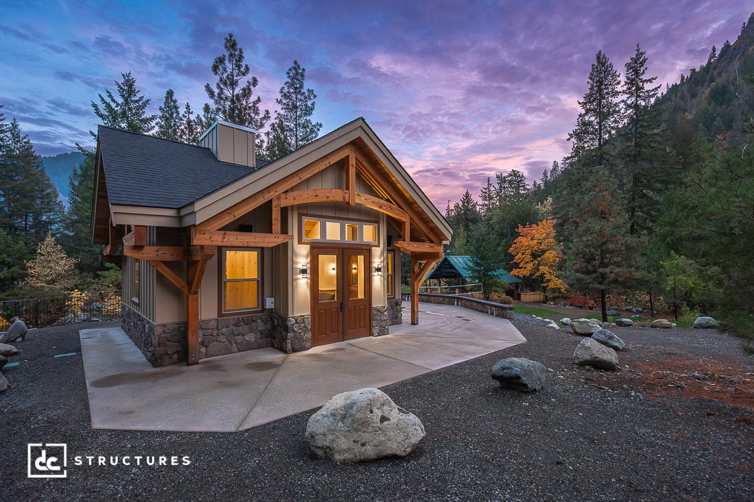 A cozy modern cabin with wooden beams and stone accents sits among trees at dusk, under a purple and pink sky with mountains.