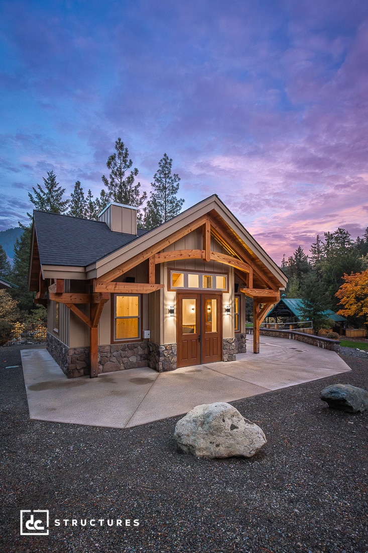 A modern cabin with stone and wood exterior, large windows, and a peaked roof stands on a patio, trees, and a colorful sunset sky.