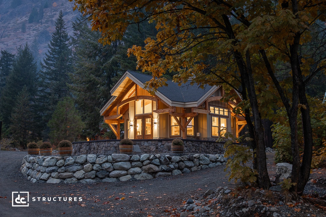 A cozy wooden cabin with large windows and warm lights sits on a stone foundation, surrounded by autumn trees at dusk.