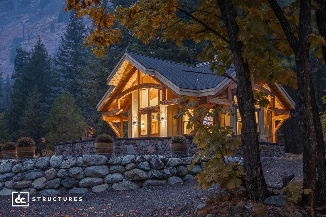 A cozy modern cabin with large windows and wooden beams sits on a stone foundation, surrounded by autumn trees and mountains at dusk. Warm lights glow inside.