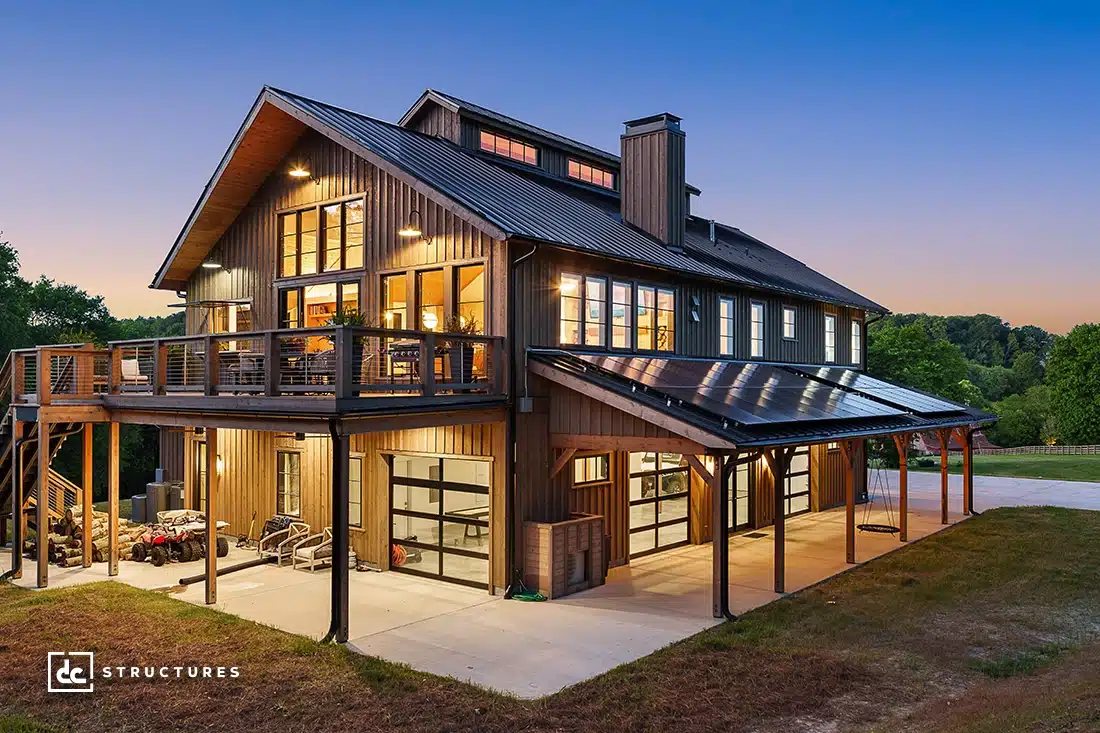 Modern two-story barn-style house with large windows, wood siding, balcony, and covered patio at sunset; warm lights inside.