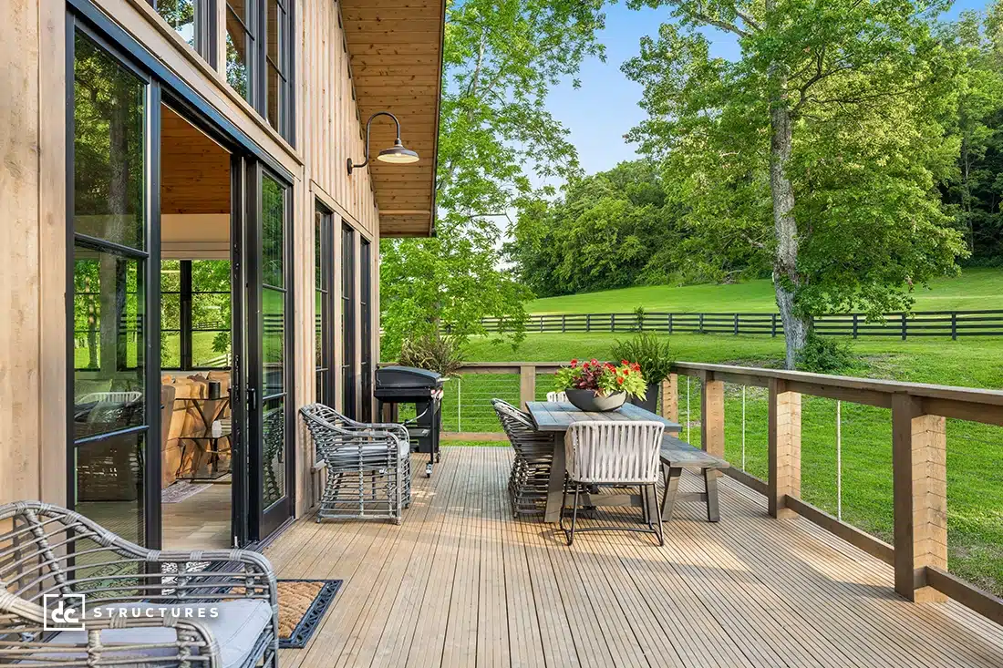 A wooden deck with wicker chairs and a dining table overlooks a green lawn and fenced pasture, with trees and blue sky in the background.