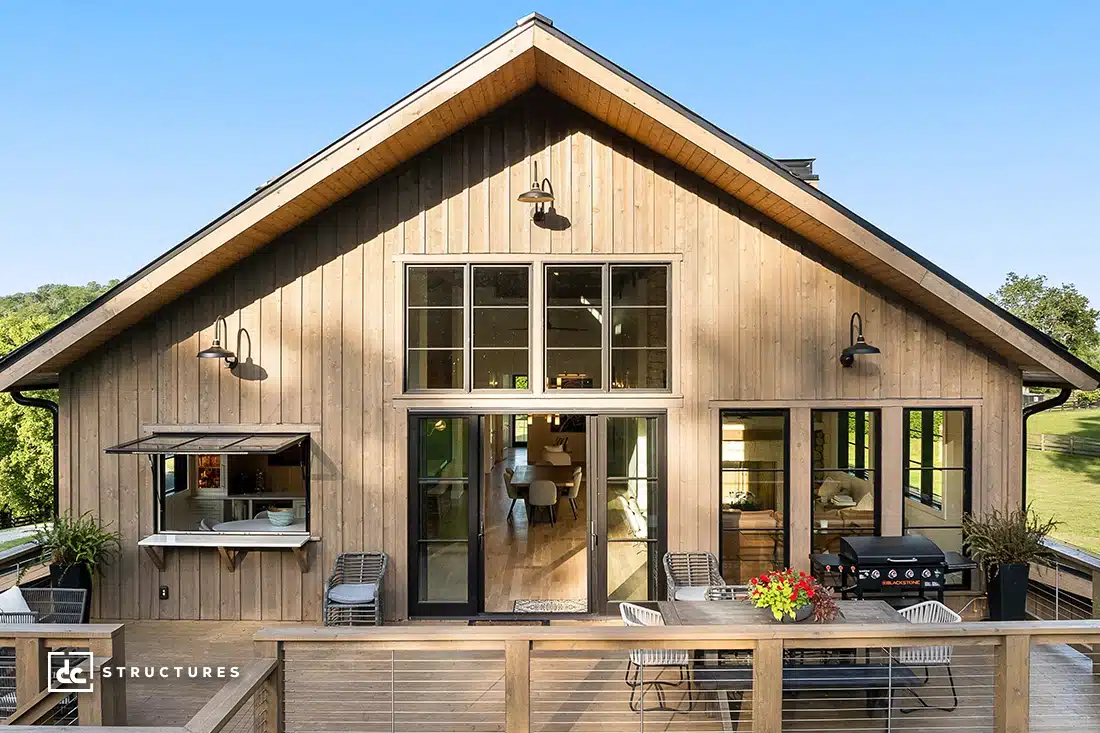 A modern wooden house with large windows and a spacious deck, featuring dining area, grill, and plants amid grass and trees.