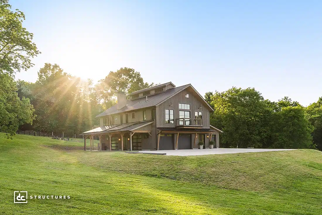 A modern two-story barn-style house with large windows sits on a grassy hill, surrounded by trees and sunlight in the background.