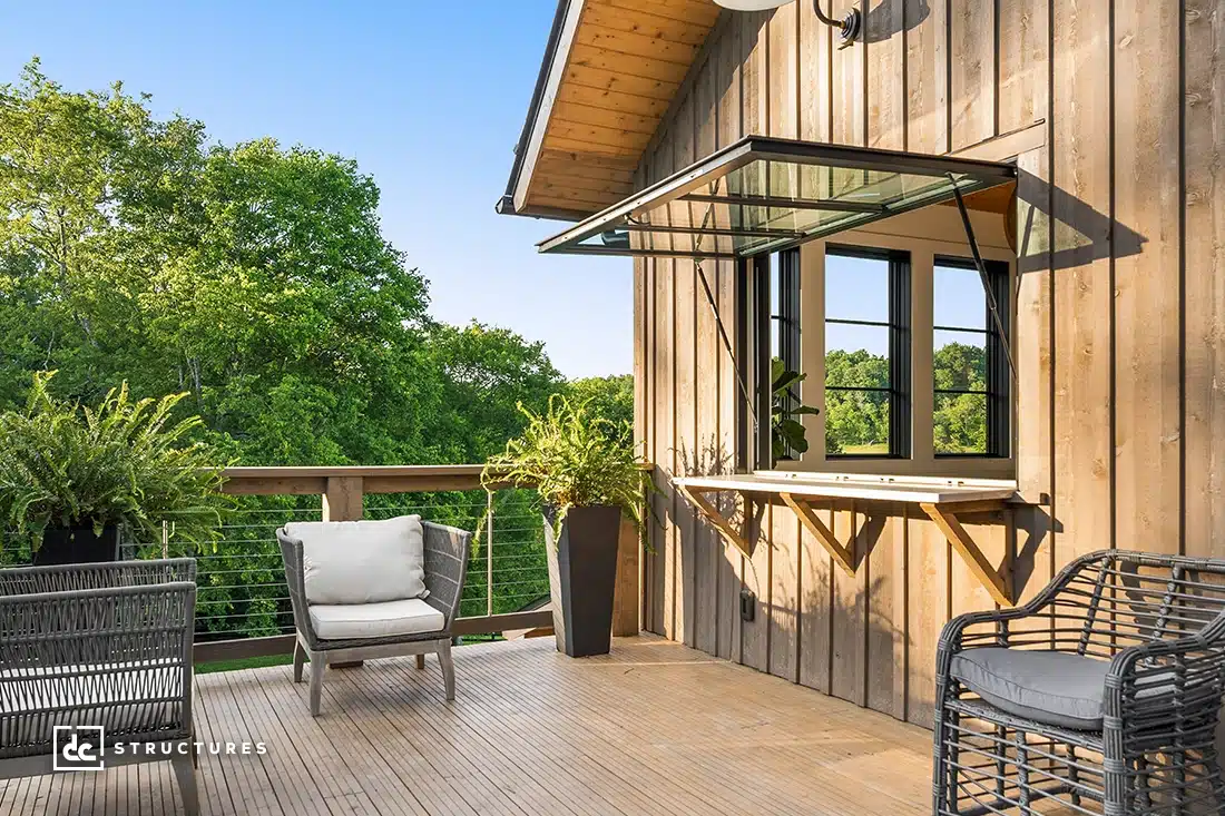 A wooden deck with modern outdoor seating, potted plants, and a large awning window overlooks green trees under a clear blue sky.