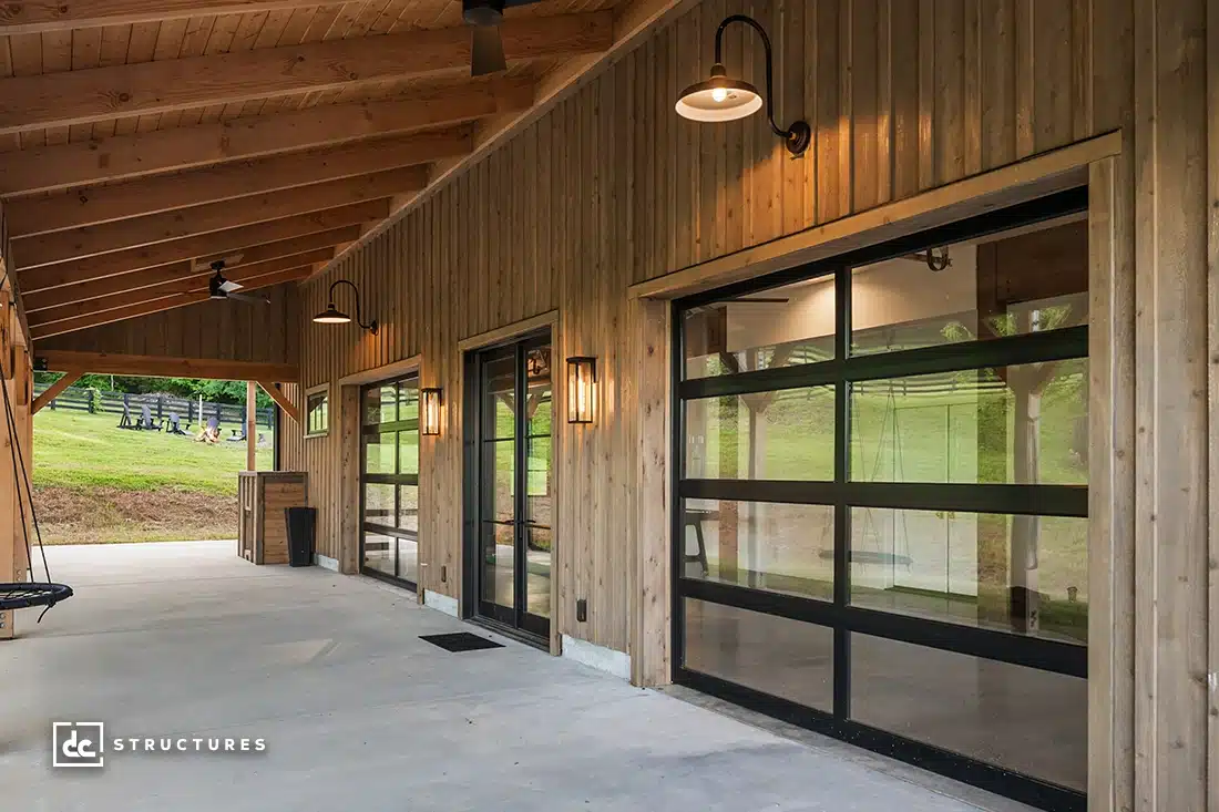A covered patio with wooden siding, exposed beams, glass garage doors, modern lantern lights, and a black porch swing overlooking grass.