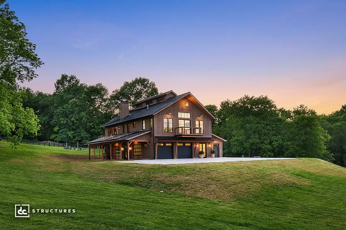 A modern two-story barn-style house with large windows and a wraparound porch, surrounded by green grass and trees at sunset.