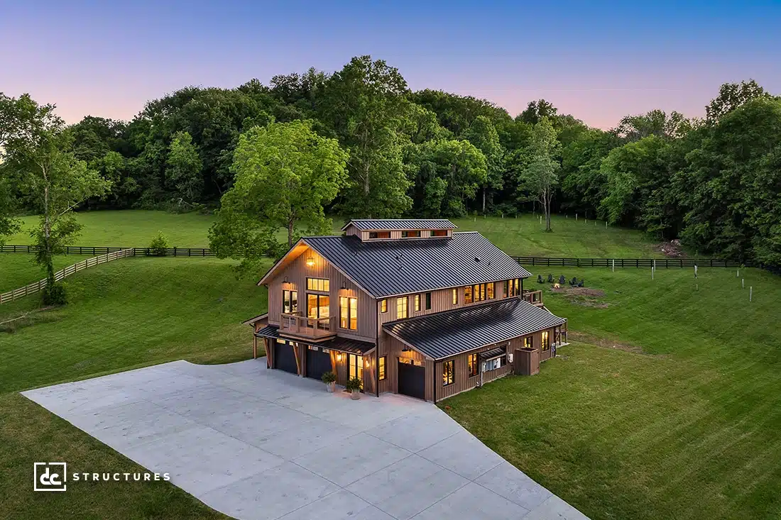 A modern two-story barn-style house with large windows sits on a green lawn surrounded by trees and a wooden fence at sunset.