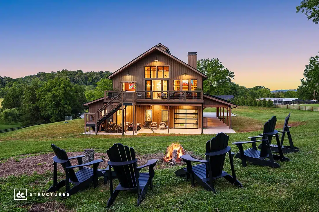 A modern two-story barn-style house with large windows and a wraparound porch, green lawn, trees, and six chairs around a fire pit at sunset.