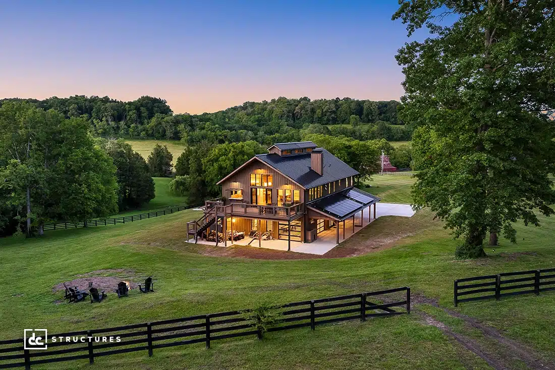 A modern two-story barn-style home with large windows is brightly lit at dusk, surrounded by grass, trees, and a wooden fence.