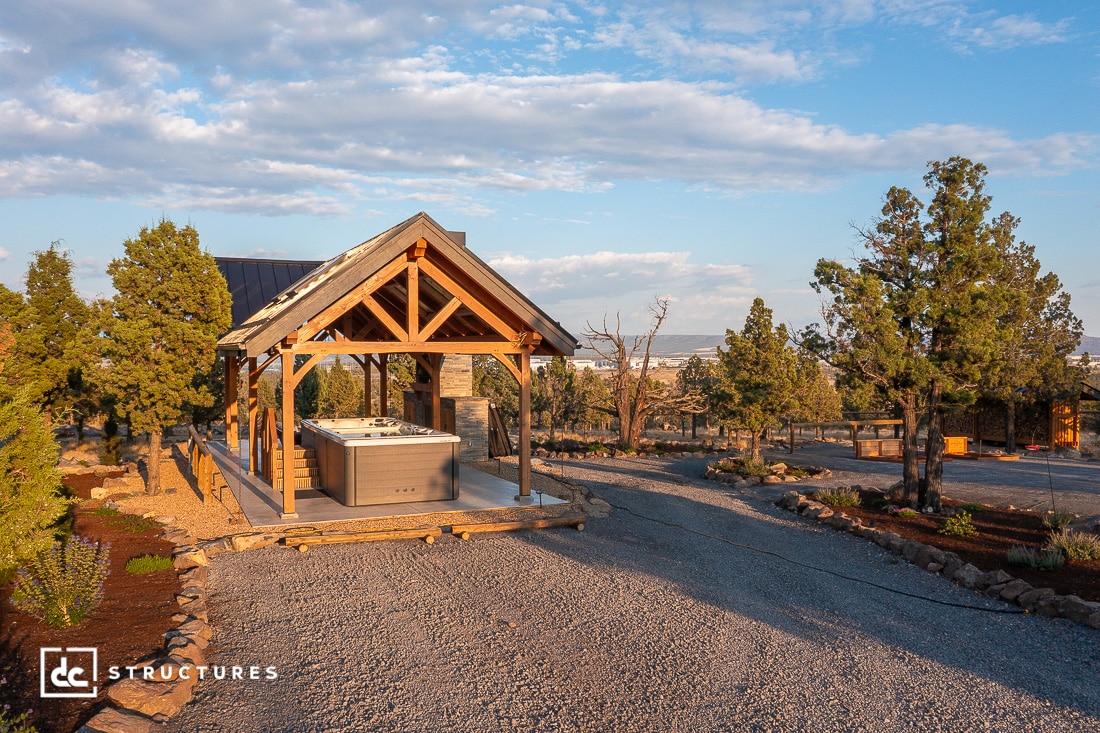 A wooden pavilion with a hot tub underneath sits on a gravel path surrounded by trees and open landscape under a partly cloudy sky.